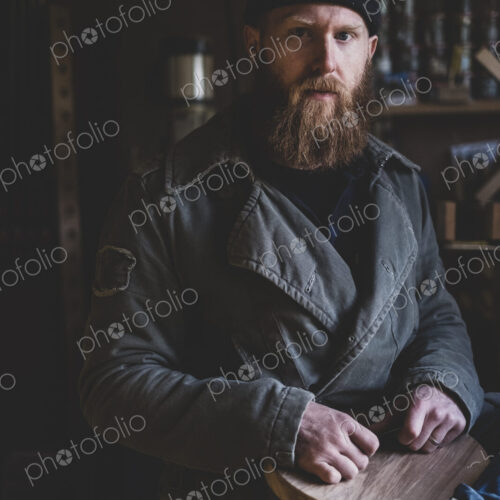 Bearded man wearing black beanie sitting in workshop, holding piece of wood, looking…