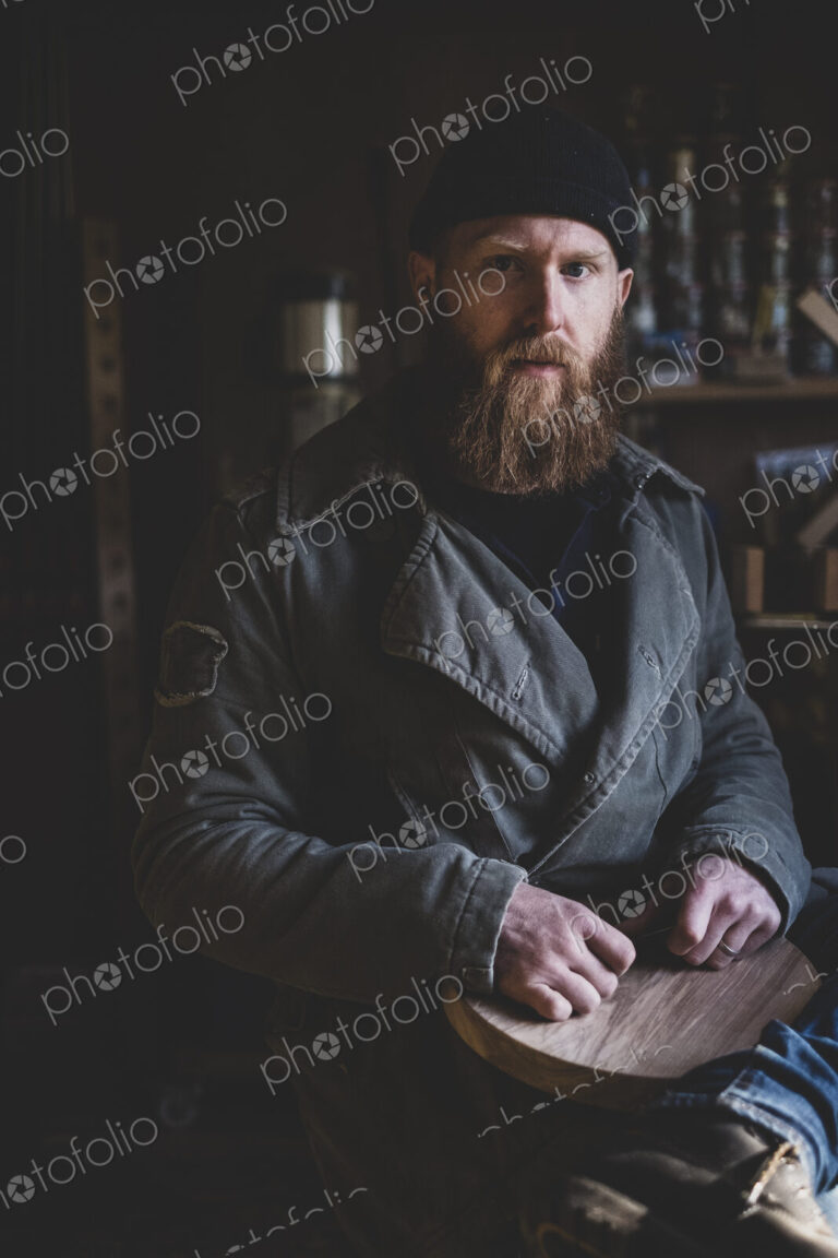Bearded man wearing black beanie sitting in workshop, holding piece of wood, looking…