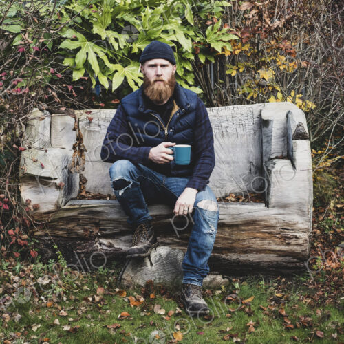 Bearded man wearing black beanie sitting on wooden bench in garden, holding blue mug,…