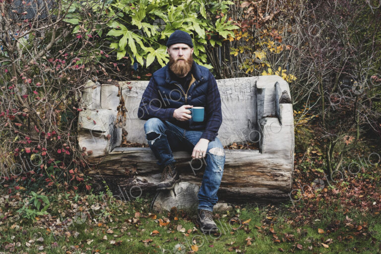 Bearded man wearing black beanie sitting on wooden bench in garden, holding blue mug,…