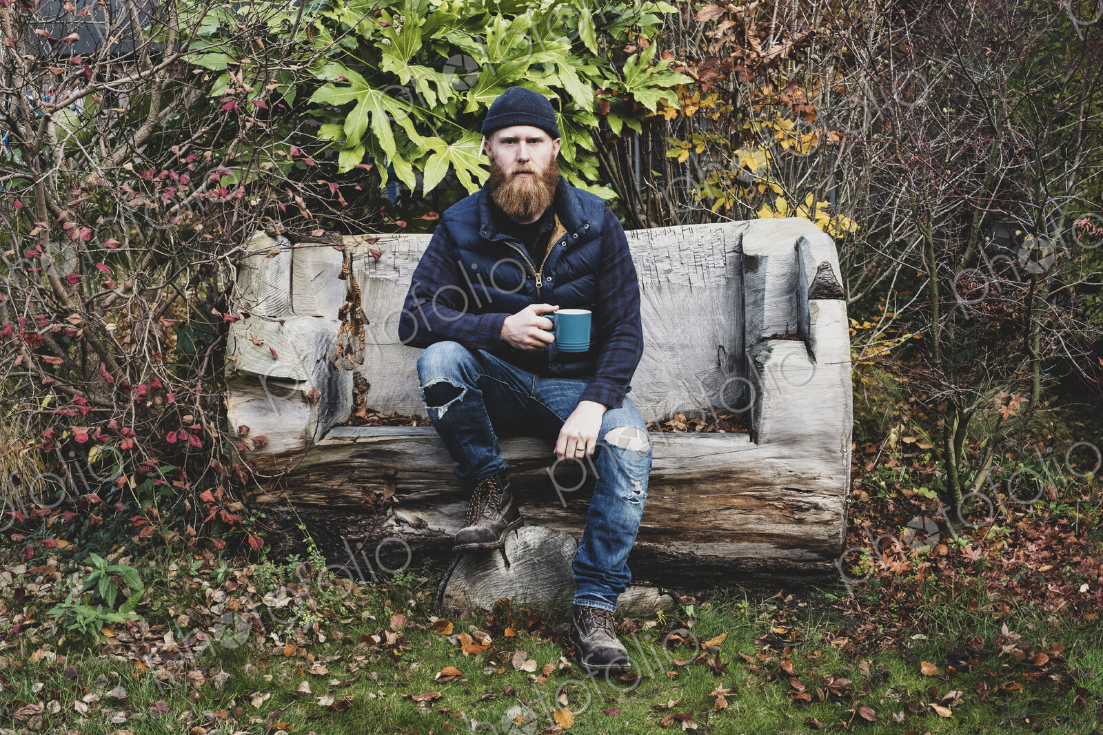 Bearded man wearing black beanie sitting on wooden bench in garden, holding blue mug,…
