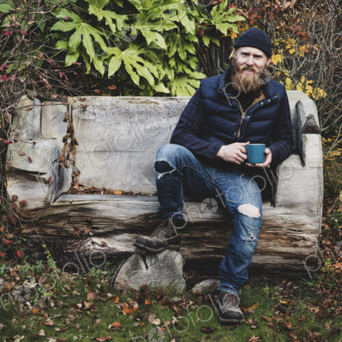 Bearded man wearing black beanie sitting on wooden bench in garden, holding blue mug,…