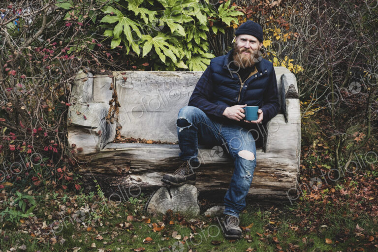 Bearded man wearing black beanie sitting on wooden bench in garden, holding blue mug,…