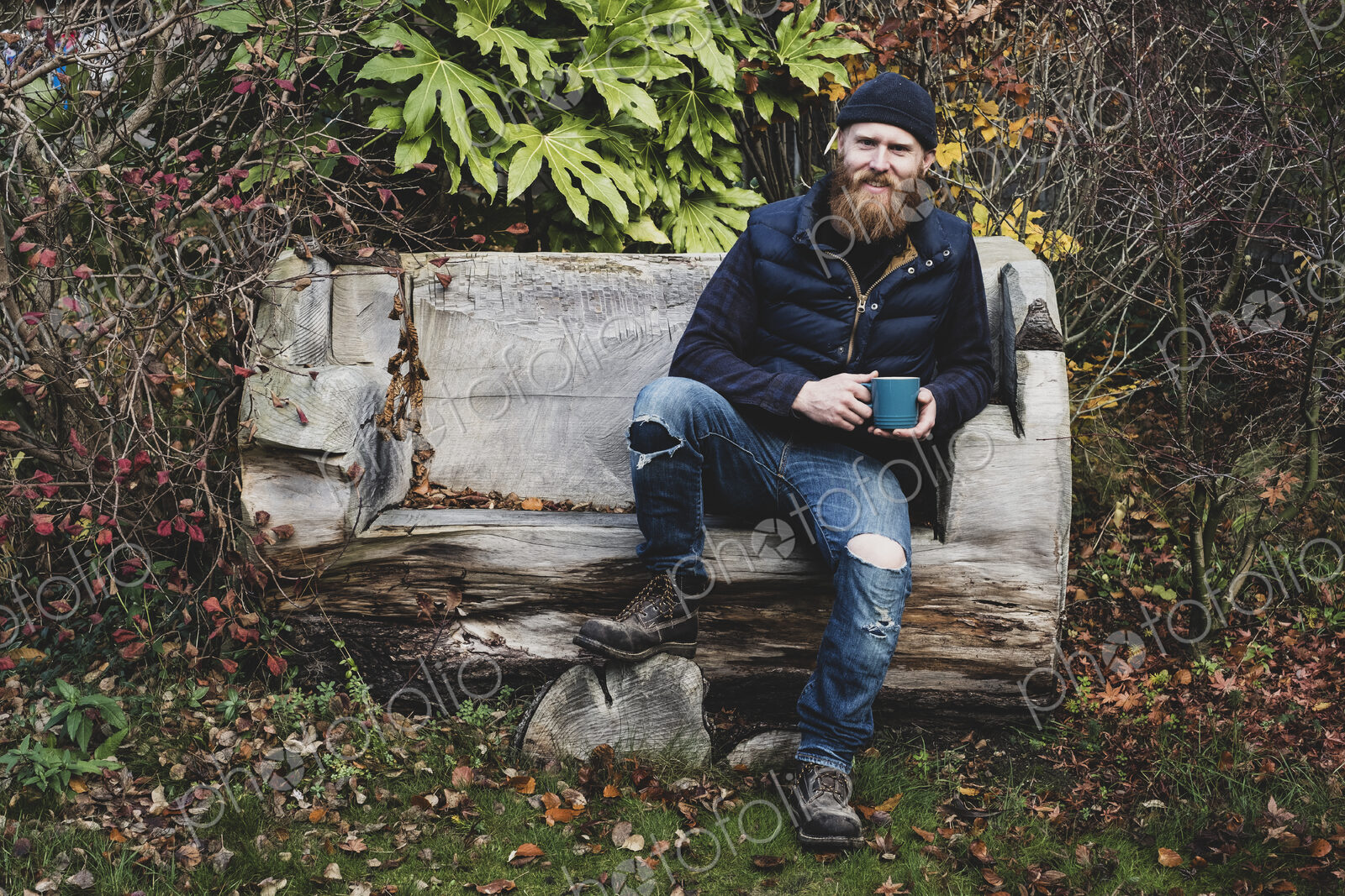 Bearded man wearing black beanie sitting on wooden bench in garden, holding blue mug,…