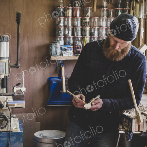 Bearded man wearing black beanie standing at workbench in workshop, examining piece of…