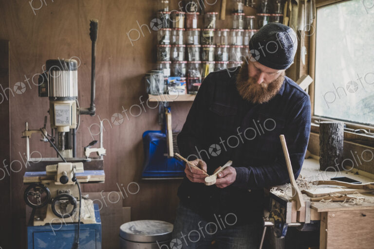Bearded man wearing black beanie standing at workbench in workshop, examining piece of…