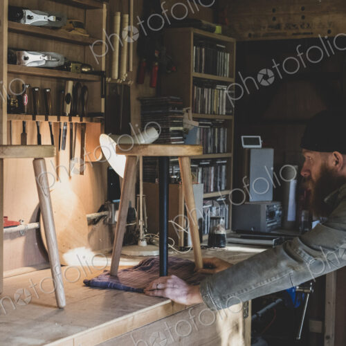 Bearded man wearing black beanie standing at workbench in workshop, examining wooden…