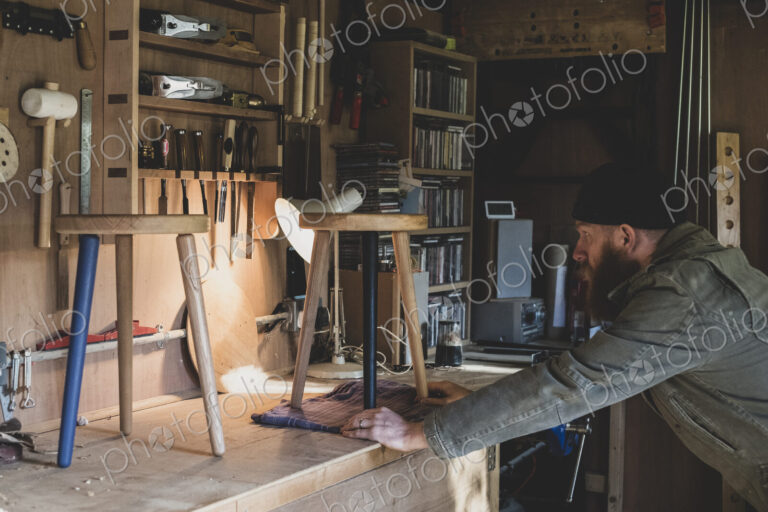Bearded man wearing black beanie standing at workbench in workshop, examining wooden…