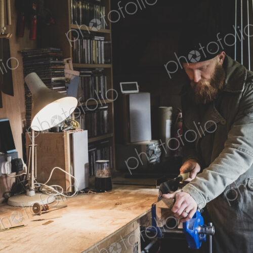 Bearded man wearing black beanie standing at workbench in workshop, sawing piece of wood. Bearded man wearing black beanie standing at workbench in workshop, sawing piece of wood.