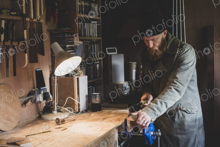 Bearded man wearing black beanie standing at workbench in workshop, sawing piece of wood.