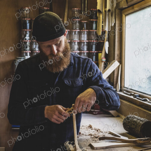 Bearded man wearing black beanie standing at workbench in workshop, working on piece… Bearded man wearing black beanie standing at workbench in workshop, working on piece…