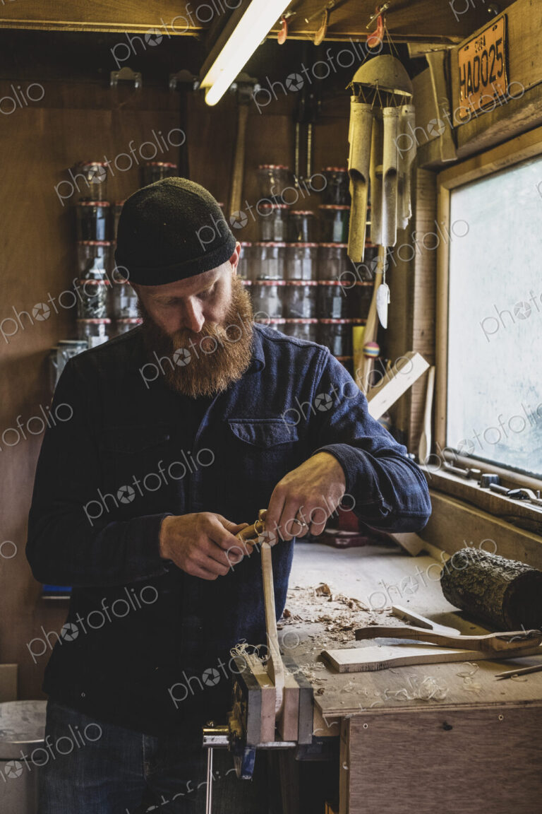 Bearded man wearing black beanie standing at workbench in workshop, working on piece…