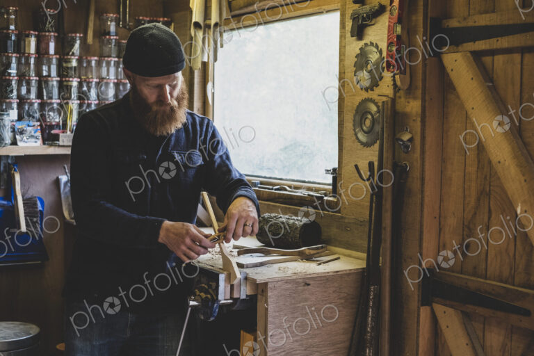 Bearded man wearing black beanie standing at workbench in workshop, working on piece…
