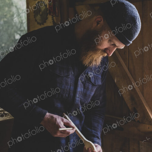 Bearded man wearing black beanie standing in workshop, examining piece of wood.