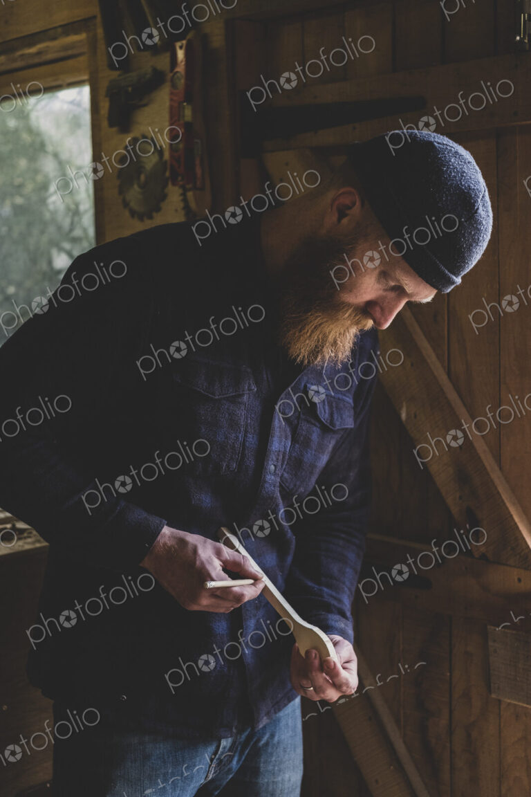 Bearded man wearing black beanie standing in workshop, examining piece of wood.