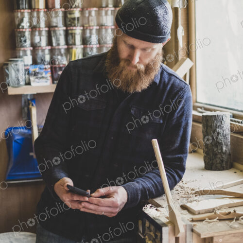 Bearded man wearing black beanie standing next to workbench in workshop, checking his…