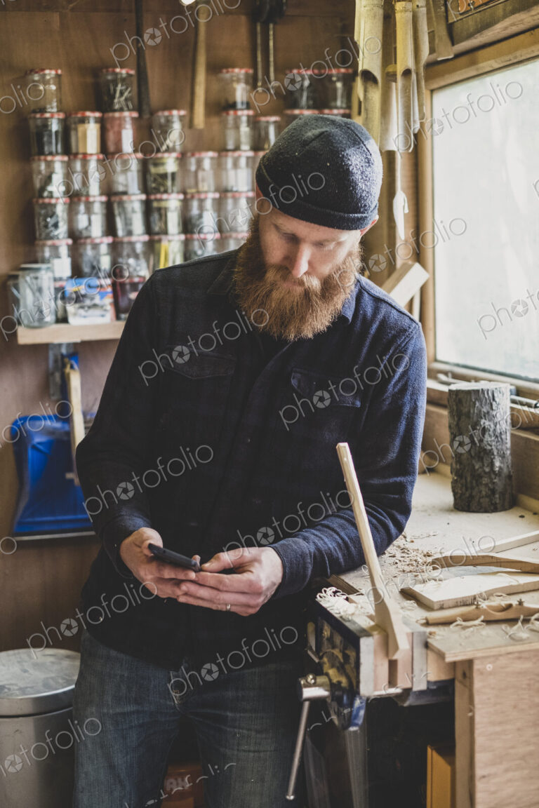 Bearded man wearing black beanie standing next to workbench in workshop, checking his…
