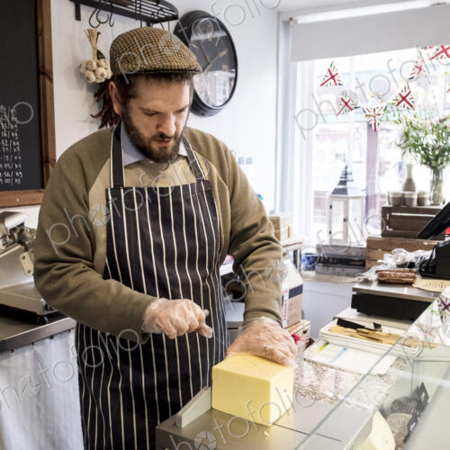 Bearded man wearing flat cap and apron standing at counter in a delicatessen, cutting… Bearded man wearing flat cap and apron standing at counter in a delicatessen, cutting…