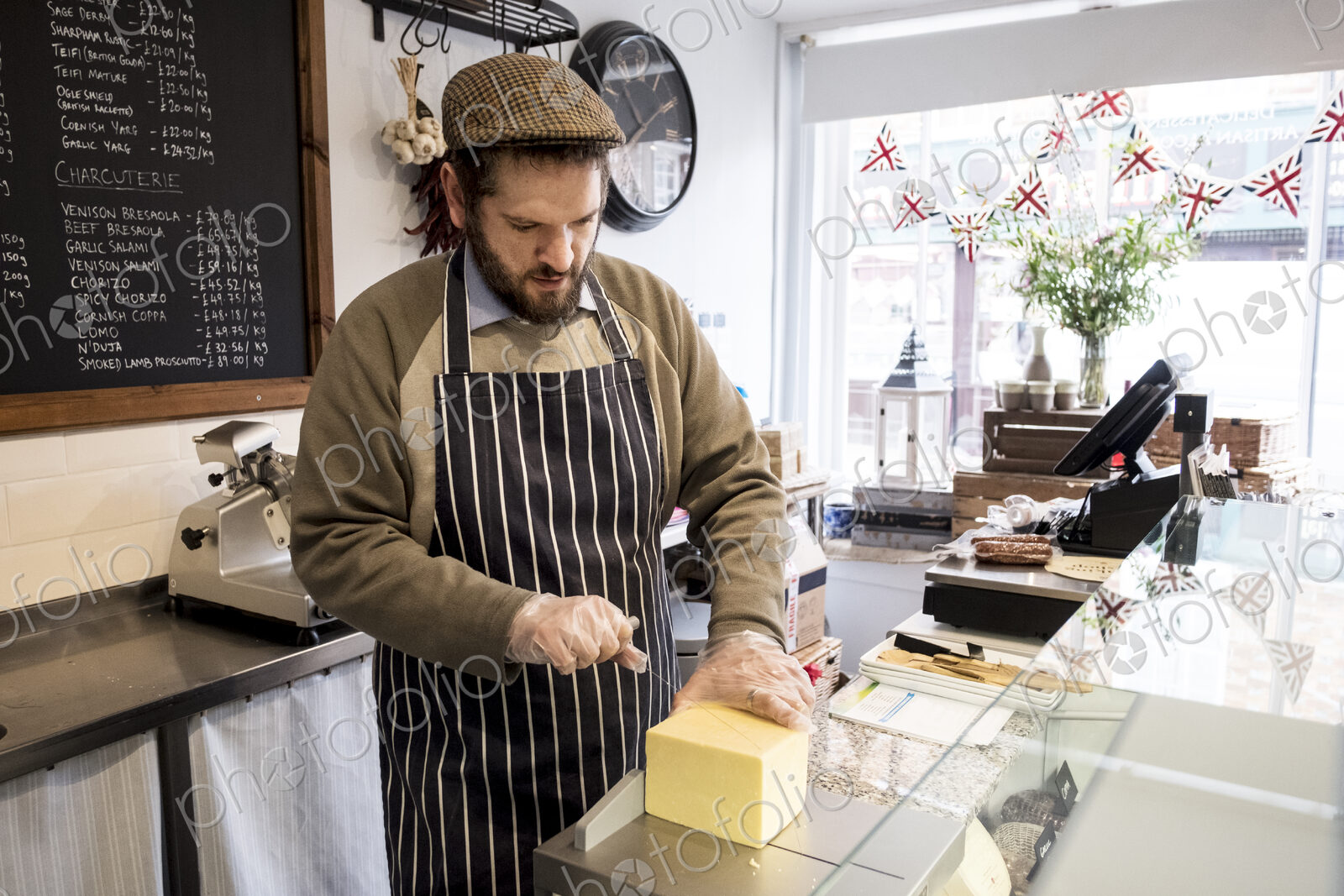Bearded man wearing flat cap and apron standing at counter in a delicatessen, cutting…