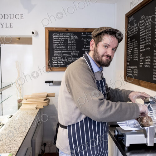 Bearded man wearing flat cap and apron standing at counter in a delicatessen, looking… Bearded man wearing flat cap and apron standing at counter in a delicatessen, looking…