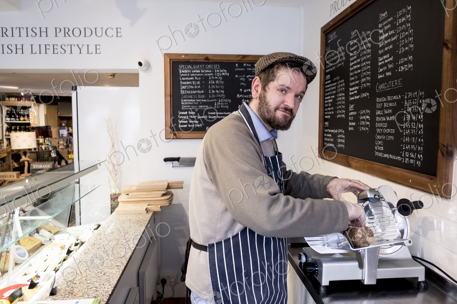 Bearded man wearing flat cap and apron standing at counter in a delicatessen, looking…
