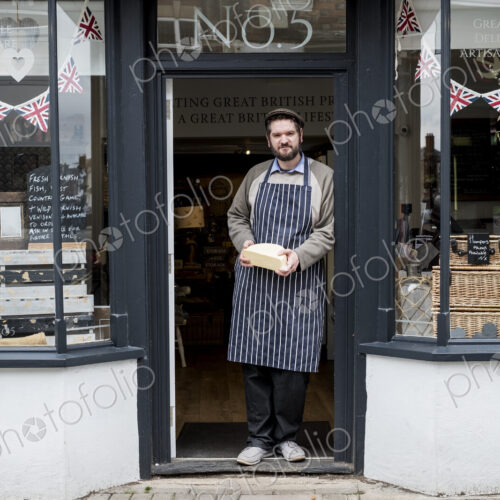 Bearded man wearing flat cap and apron standing in doorway of a delicatessen, holding… Bearded man wearing flat cap and apron standing in doorway of a delicatessen, holding…