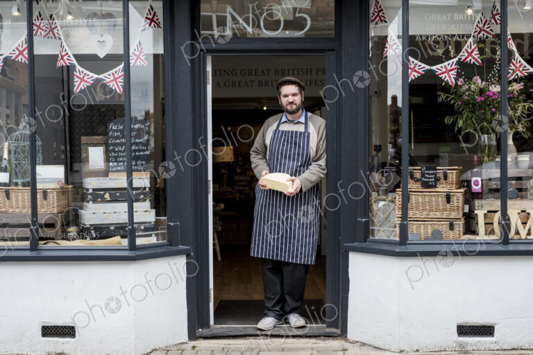 Bearded man wearing flat cap and apron standing in doorway of a delicatessen, holding…