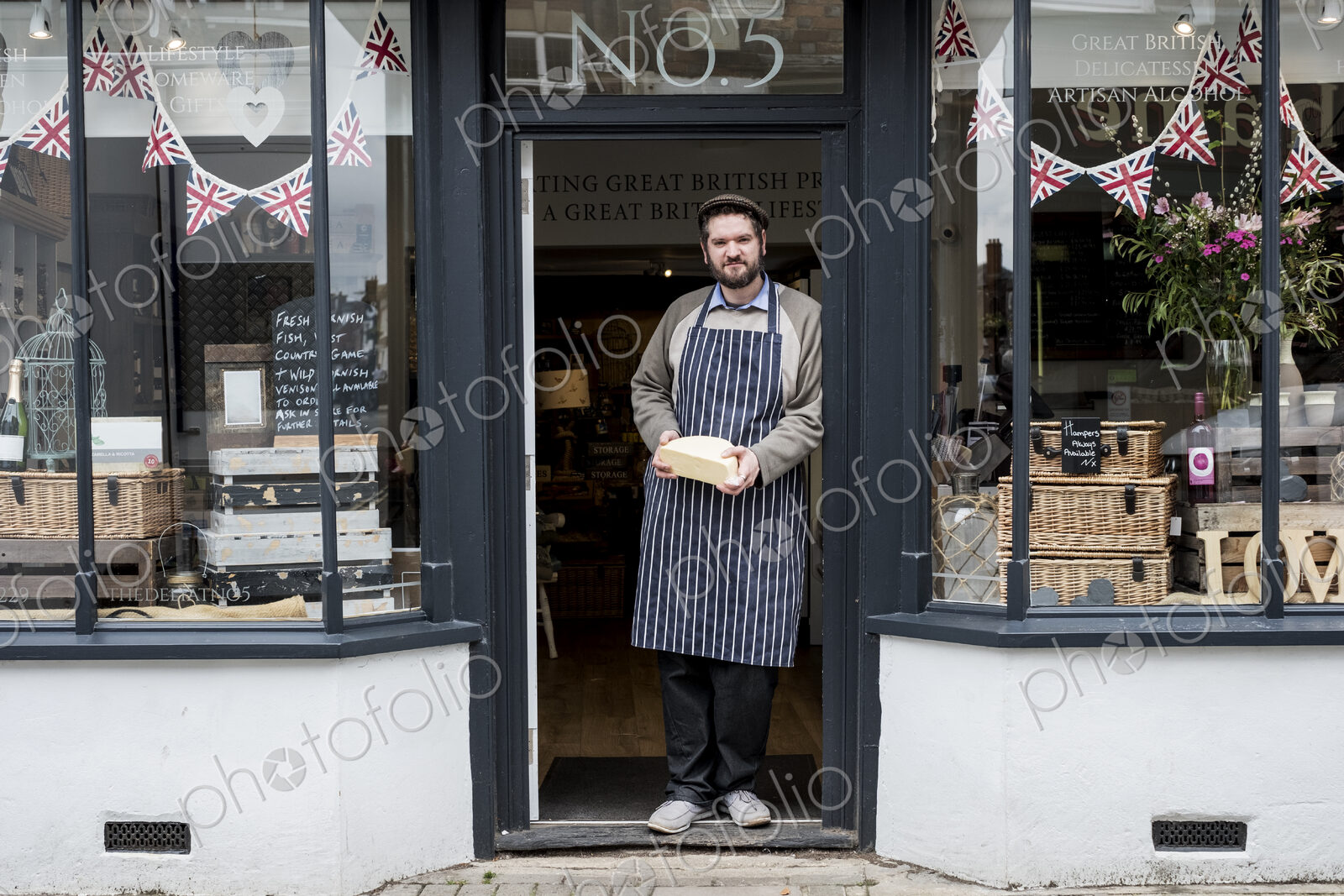 Bearded man wearing flat cap and apron standing in doorway of a delicatessen, holding…
