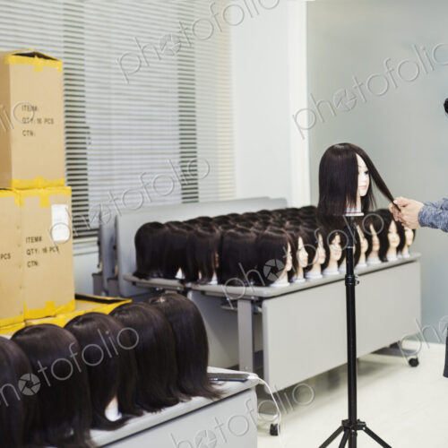 Bearded man wearing glasses standing indoors, cutting hair of brown wig on mannequin head.