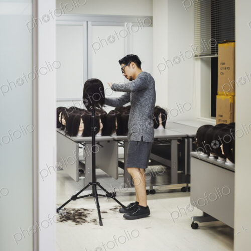 Bearded man wearing glasses standing indoors, cutting hair of brown wig on mannequin head.