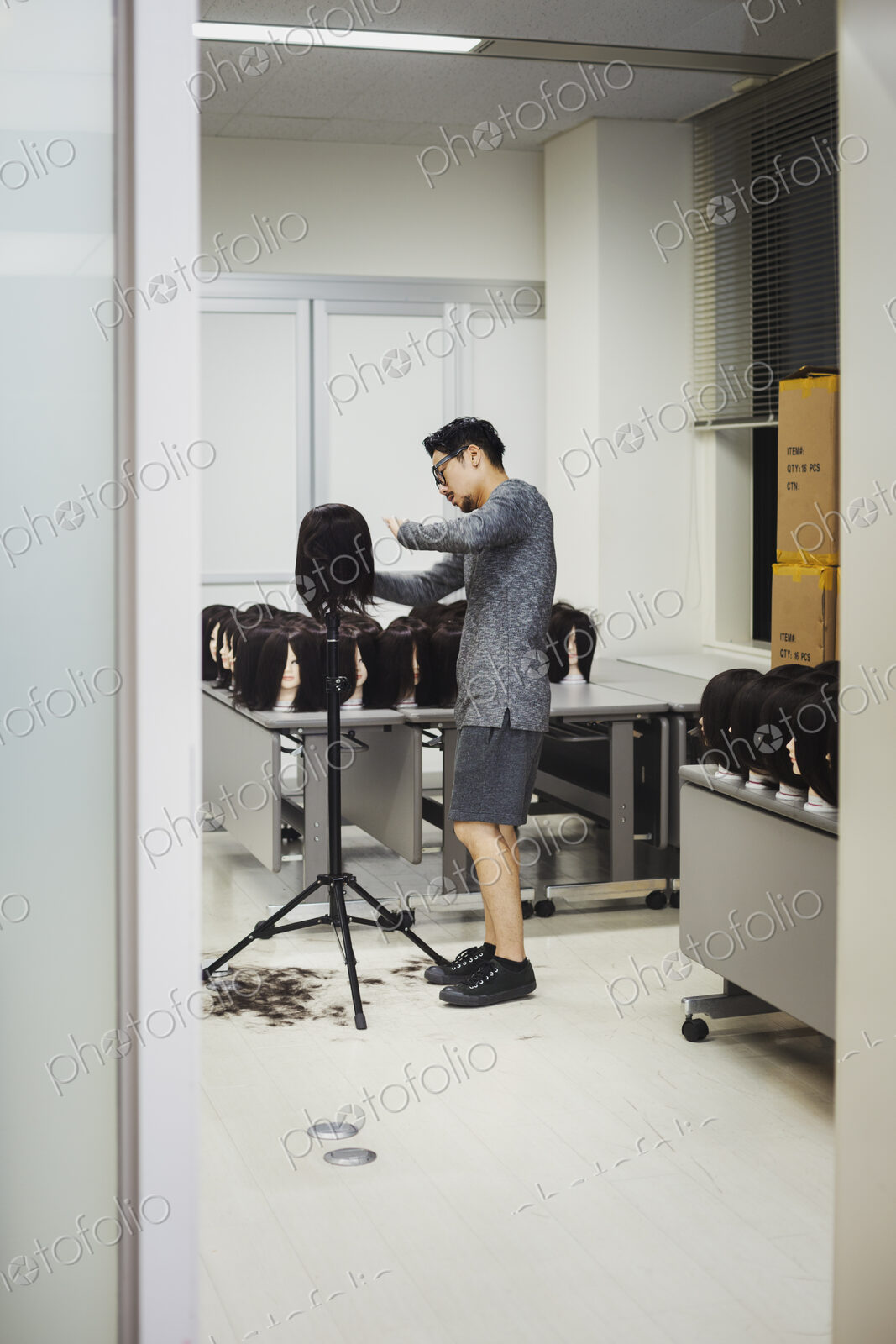 Bearded man wearing glasses standing indoors, cutting hair of brown wig on mannequin head.