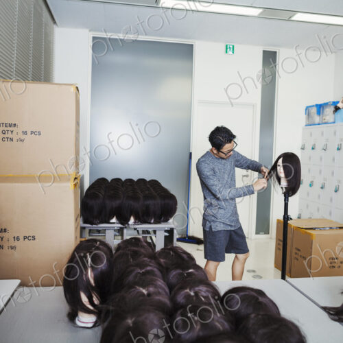 Bearded man wearing glasses standing indoors, cutting hair of brown wig on mannequin head.