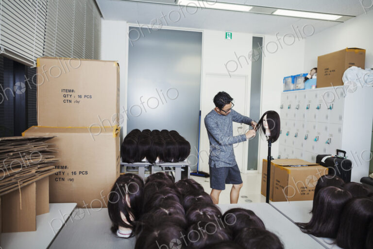 Bearded man wearing glasses standing indoors, cutting hair of brown wig on mannequin head.