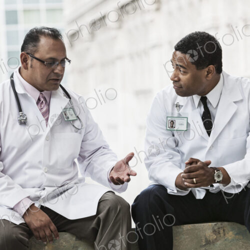 Black and Hispanic male doctors discussing a case in a hospital hallway. Black and Hispanic male doctors discussing a case in a hospital hallway.