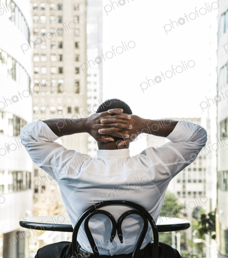 Black business man sitting at a lobby table looking at the cityscape through a large…