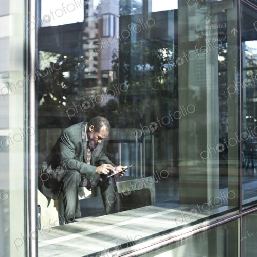 Black business man texting while sitting in front of a large widow in a lobby waiting… Black business man texting while sitting in front of a large widow in a lobby waiting…
