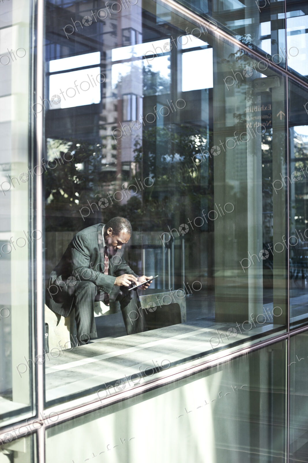 Black business man texting while sitting in front of a large widow in a lobby waiting…