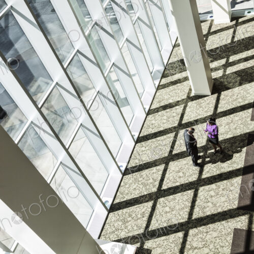Black businessman and woman meeting next to a large bank of windows in the lobby of a… Black businessman and woman meeting next to a large bank of windows in the lobby of a…