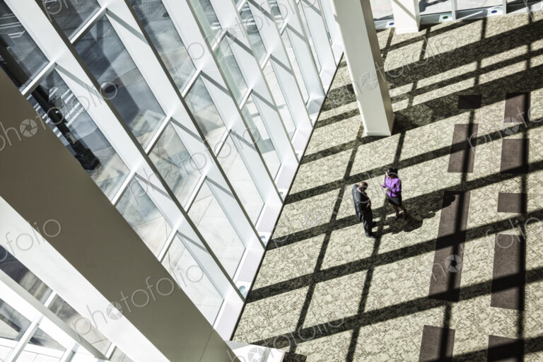 Black businessman and woman meeting next to a large bank of windows in the lobby of a…