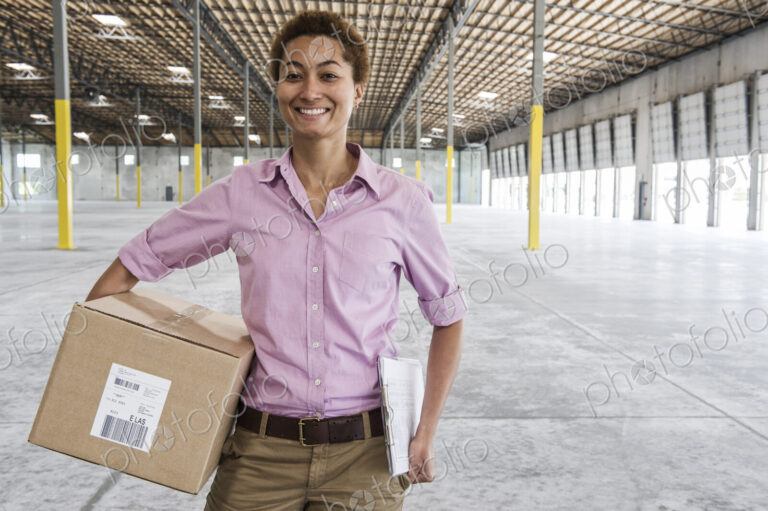 Black female holding cardboard box and standing in front of loading dock doors in a…