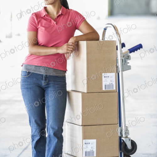 Black female standing next to hand truck in front of loading dock doors inside of a… Black female standing next to hand truck in front of loading dock doors inside of a…