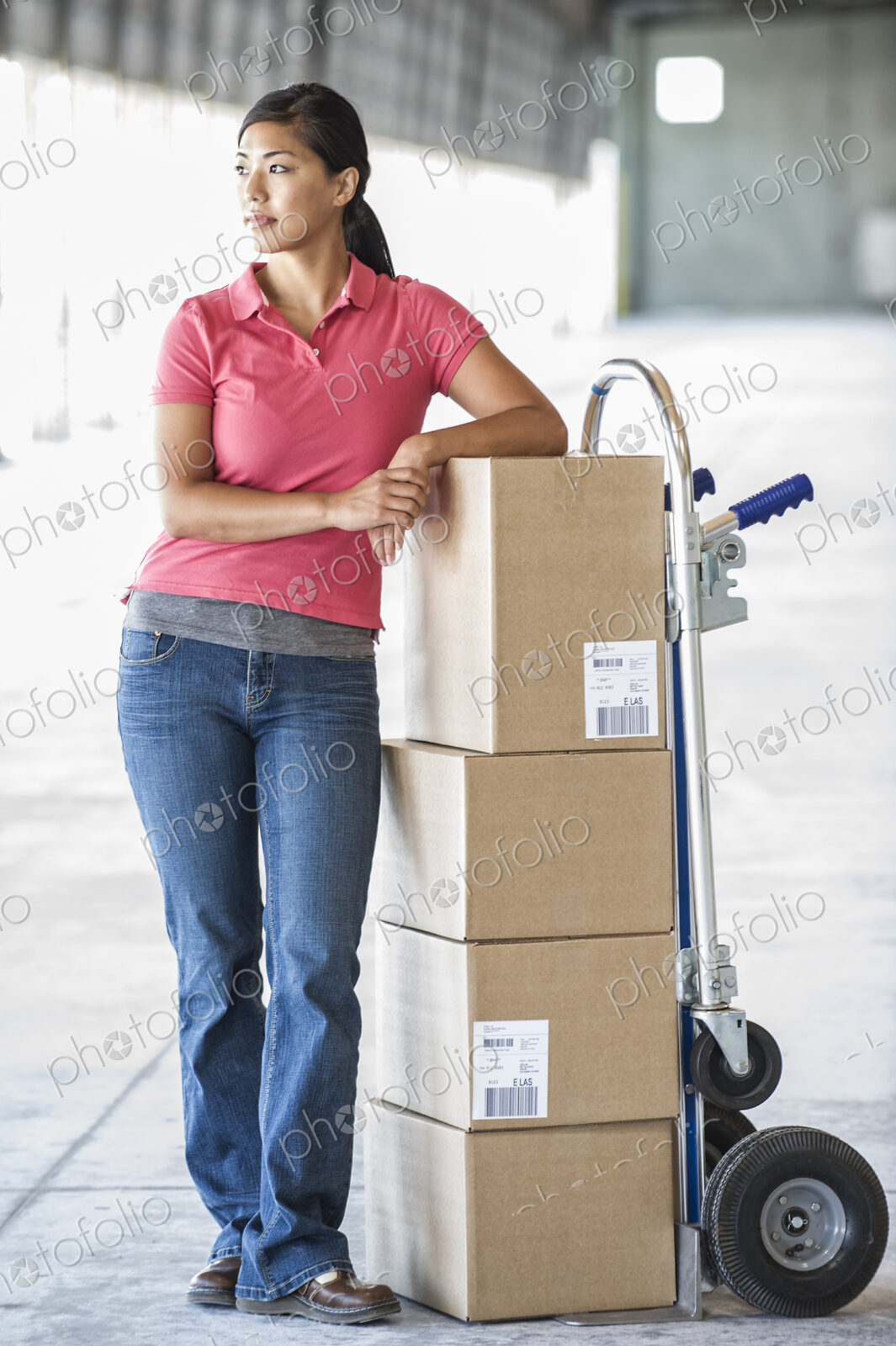 Black female standing next to hand truck in front of loading dock doors inside of a…