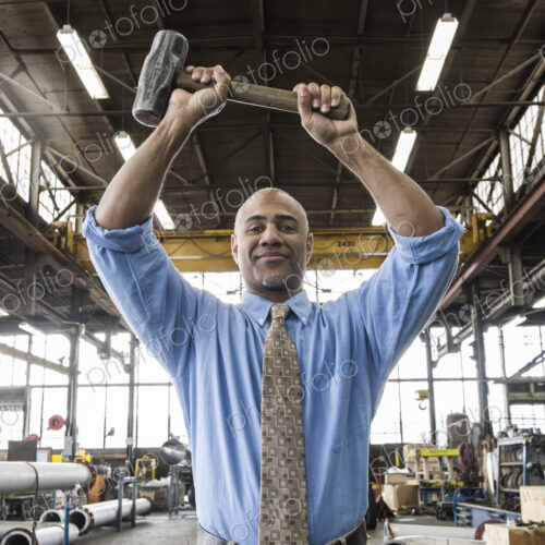 Black man owner of a sheet metal factory holding a hammer while standing on the floor…