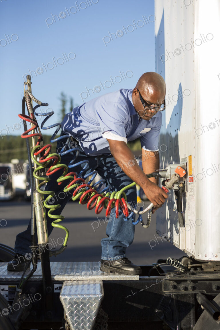 Black man truck driver attaching power cables from truck tractor to trailer at a truck…