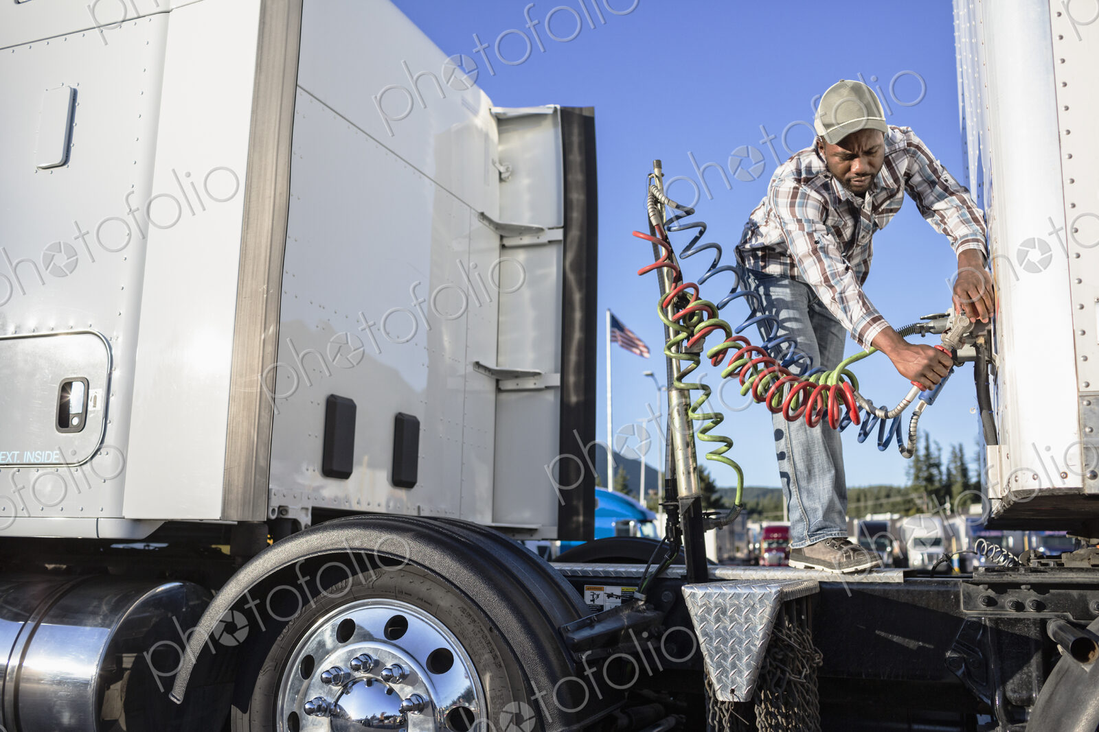 Black man truck driver attaching power cables from truck tractor to trailer at a truck…