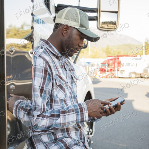 Black man truck driver texting while standing next to his truck cab parked in a lot at…