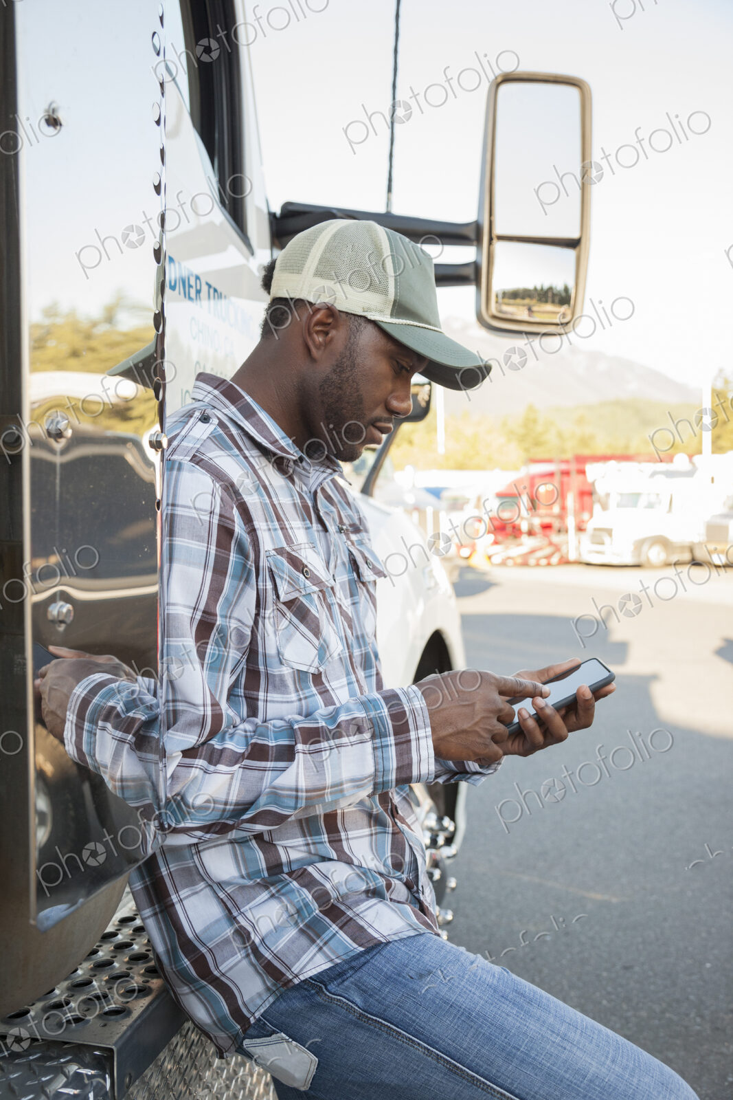 Black man truck driver texting while standing next to his truck cab parked in a lot at…