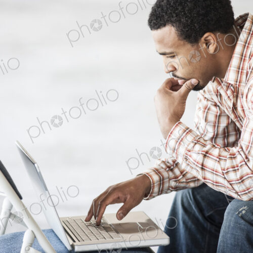 Black man working on a lap top computer and usiing a chair as a table top. Black man working on a lap top computer and usiing a chair as a table top.