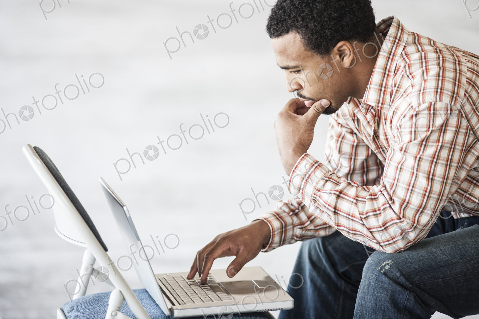 Black man working on a lap top computer and using a chair as a table top.