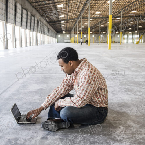 Black man working on lap top computer in front of loading dock doors in a new waerhouse. Black man working on lap top computer in front of loading dock doors in a new waerhouse.